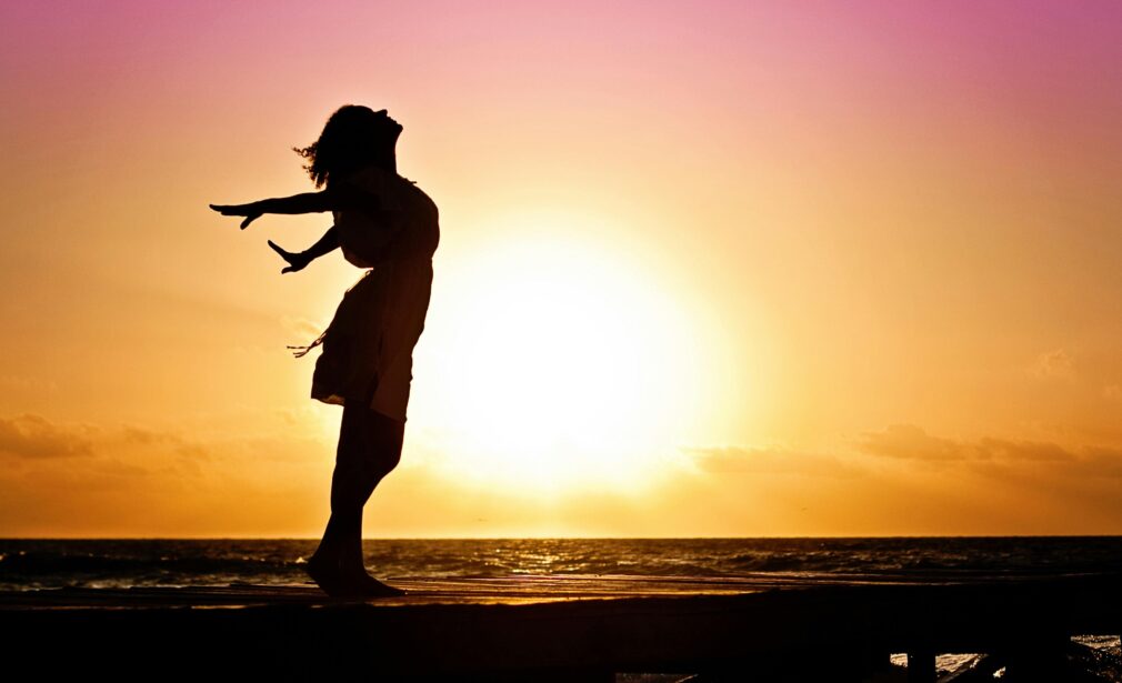 Silhouette of a woman at the beach with arms outstretched against a vibrant sunset backdrop.